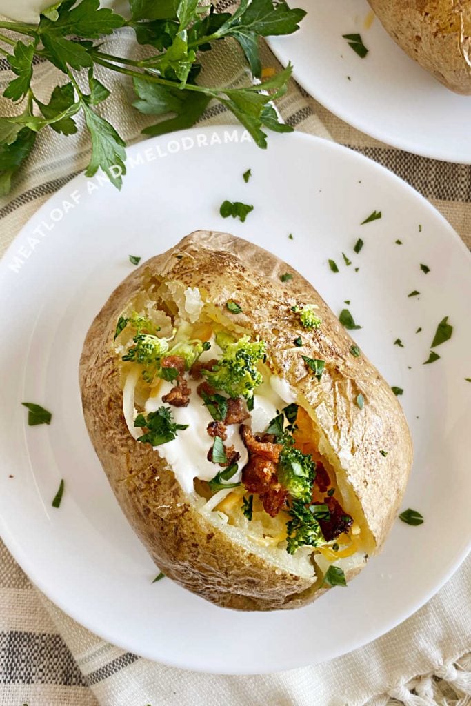 microwaved russet potato with sour cream, bacon, broccoli and cheese on white plate with parsley 