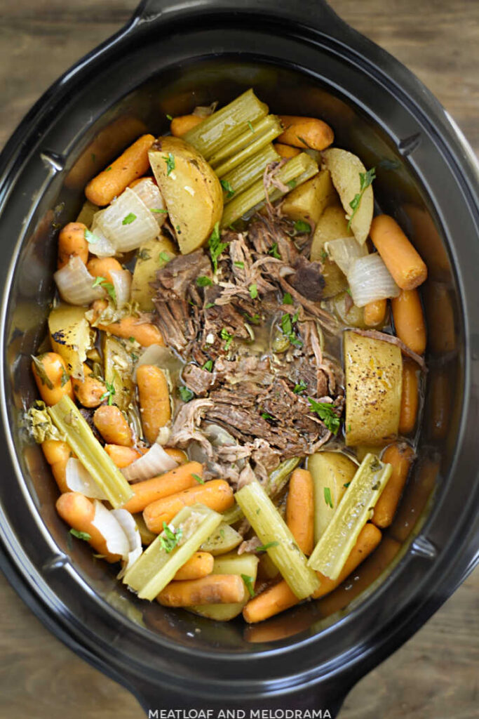 slow cooked beef chuck roast with baby carrots, cut potatoes and celery stalks topped with parsley in the crock pot