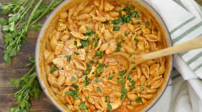 chicken and pasta shells in a creamy tomato sauce cooked in red dutch oven with wooden spoon and topped with parsley
