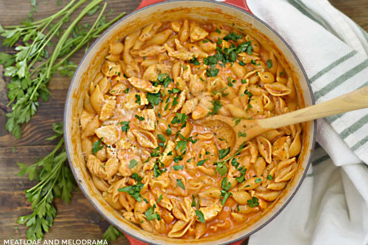 chicken and pasta shells in a creamy tomato sauce cooked in red dutch oven with wooden spoon and topped with parsley