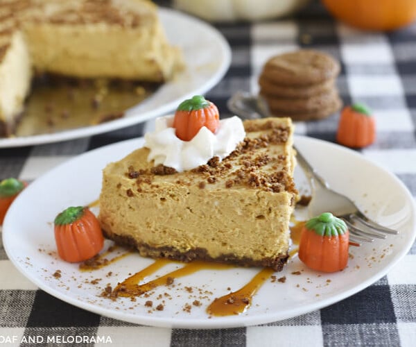 instant pot pumpkin cheesecake with gingerbread crust on a white plate with whipped cream, caramel and marshmallow pumpkins