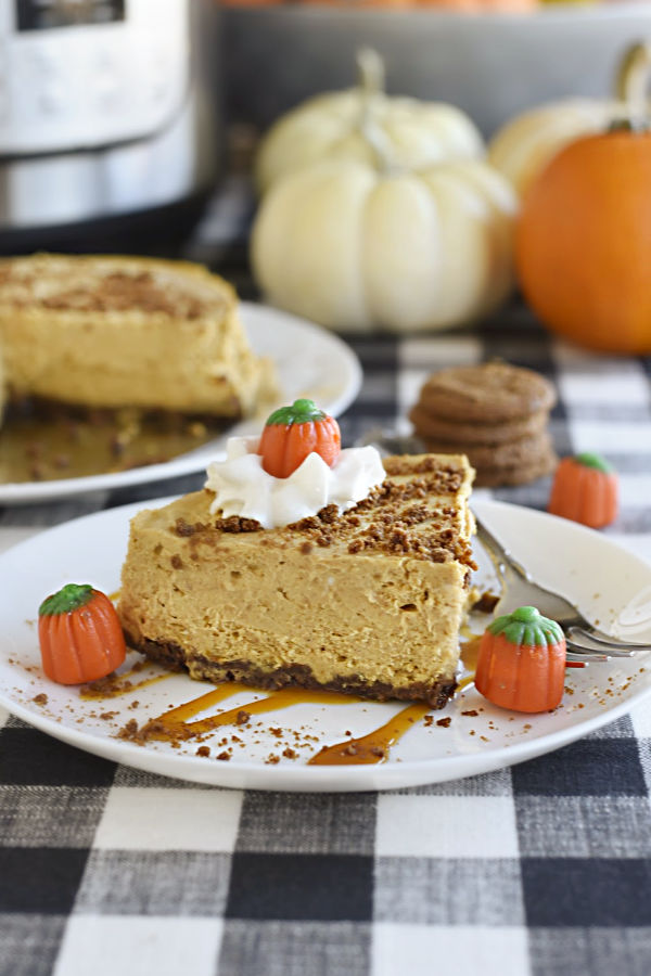 pumpkin cheesecake with gingerbread crust on a white plate over caramel topped with whipped cream, candy pumpkins with pumpkins and instant pot in background on buffalo check tablecloth