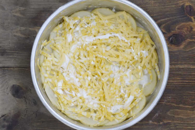 sliced potatoes layered with cream and shredded cheese in round cake pan