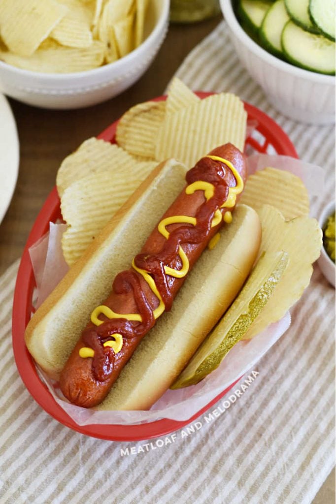hot dog with mustard, ketchup, pickle and potato chips in red dish