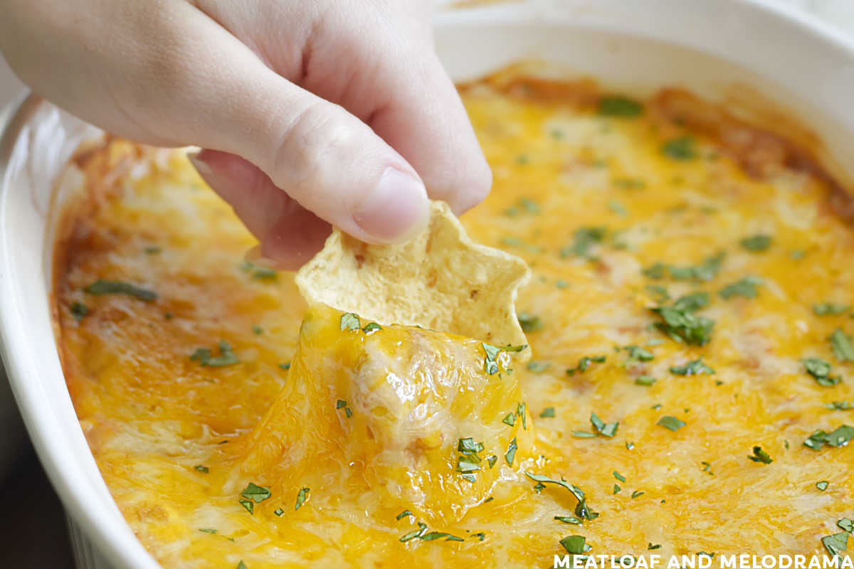 hand dipping tortilla chip into hot bean dip with melted cheese on top