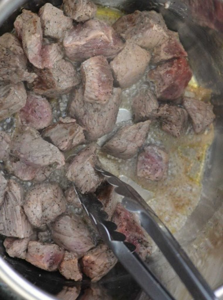 brown chunks of beef stew meat in pressure cooker on saute with kitchen tongs