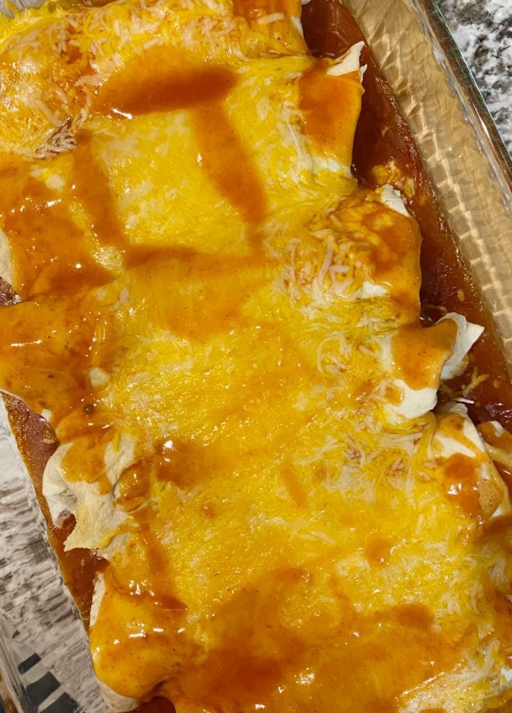 baked smothered burritos in a baking dish with red enchilada sauce and melted cheddar cheese