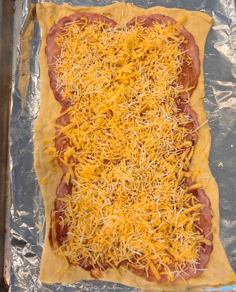 crescent dough sheet topped with ham slices and shredded cheese on baking sheet
