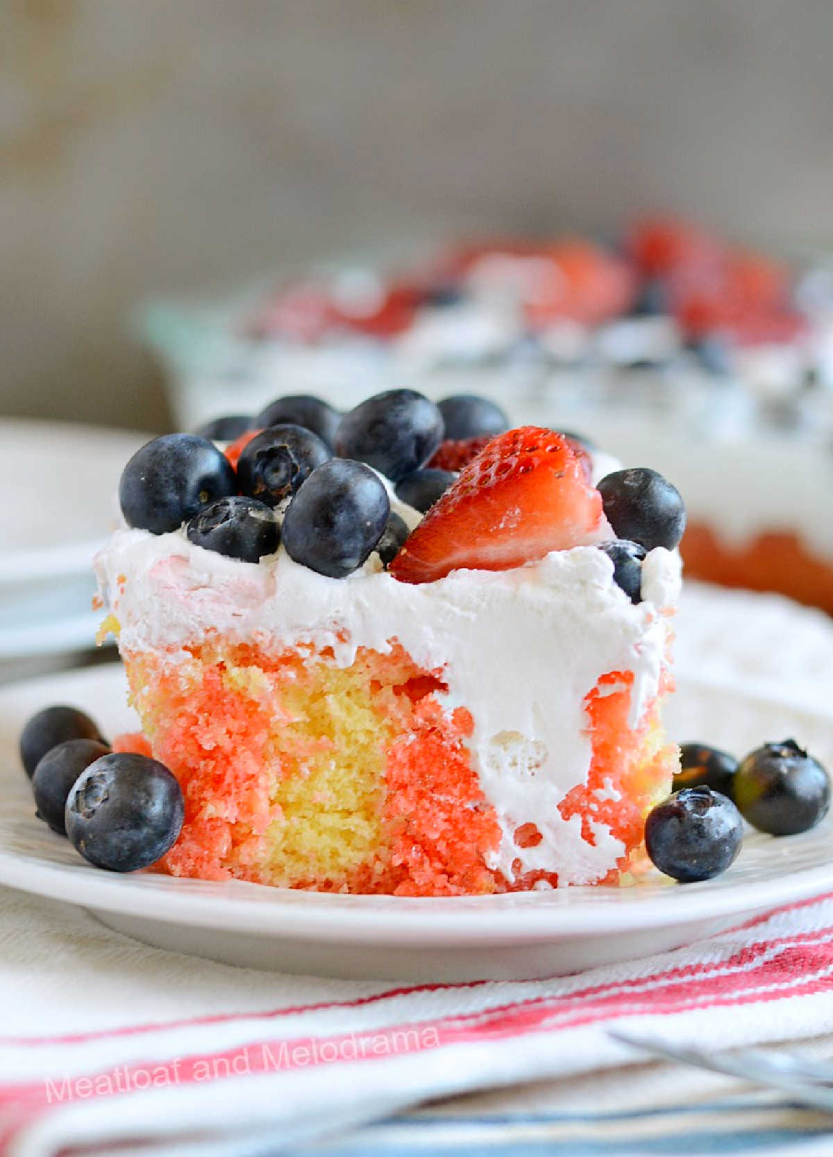 slice of red white and blue poke cake with cool whip, strawberries and blueberries on plate