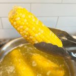 kitchen tongs holding boiled corn over pot of boiling water with butter