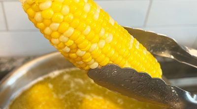kitchen tongs holding boiled corn over pot of boiling water with butter