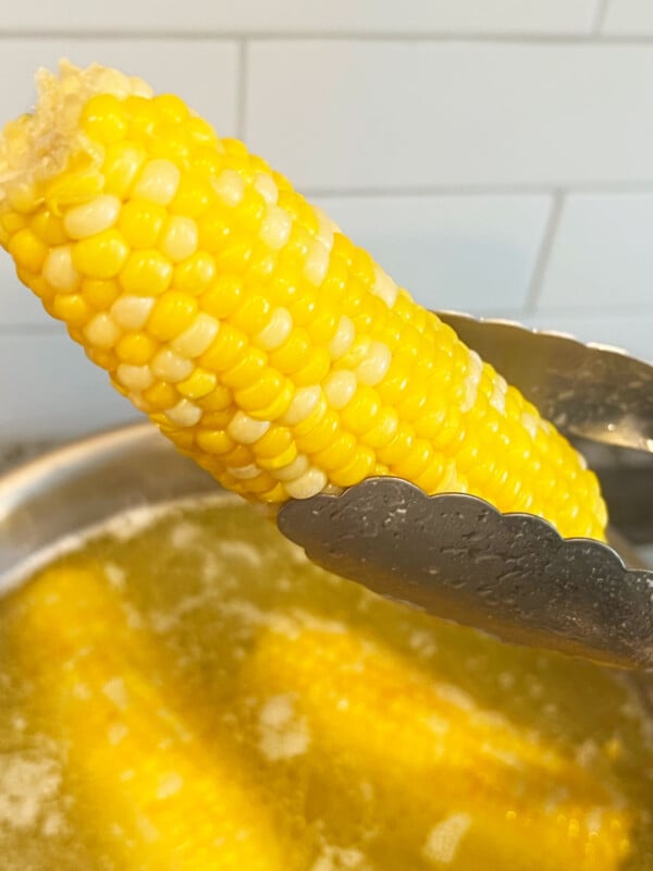 kitchen tongs holding boiled corn over pot of boiling water with butter