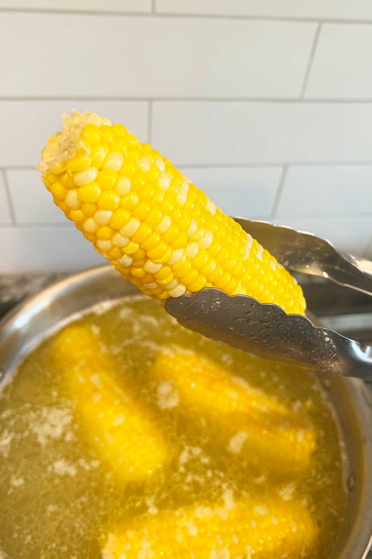 ear of boiled corn in kitchen tongs over pot of boiling water with butter