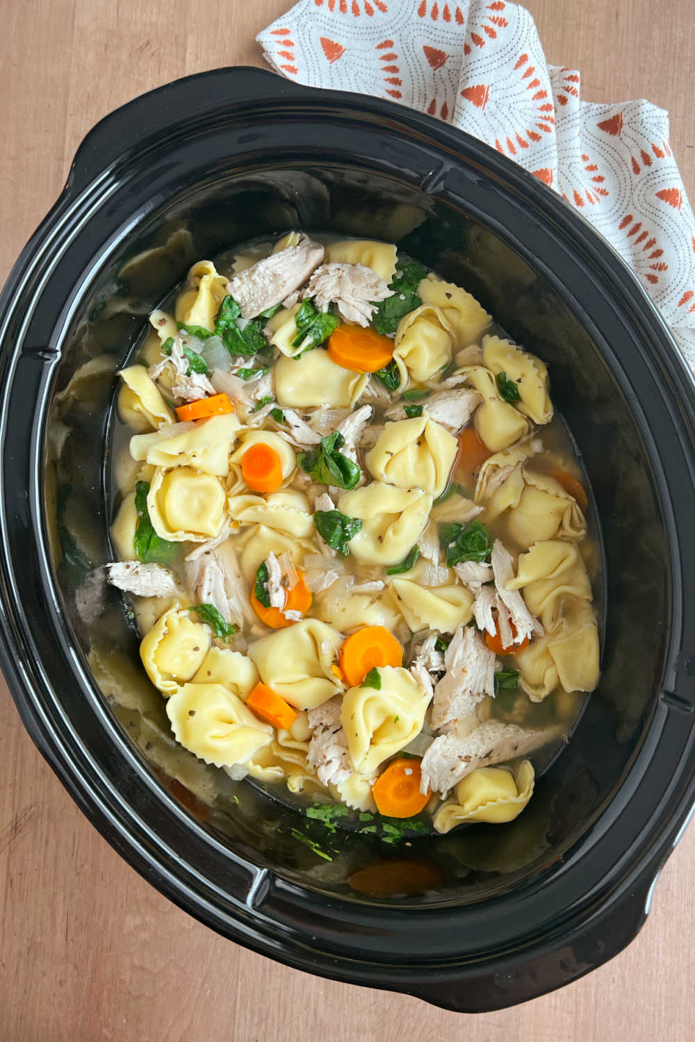 crock pot chicken tortellini soup with shredded chicken, carrots and fresh spinach in slow cooker.