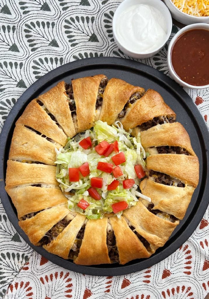 taco ring recipe with crescent roll dough and ground beef on plate with lettuce and tomatoes.