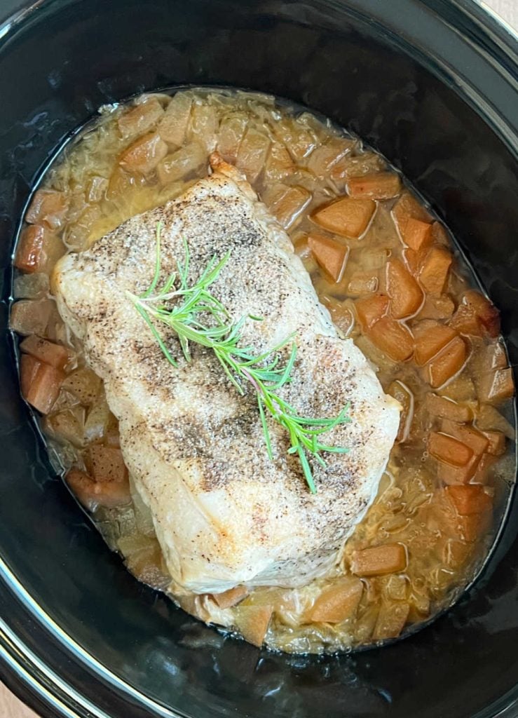 whole pork loin with rosemary sprig over apples and sauerkraut in crock pot.
