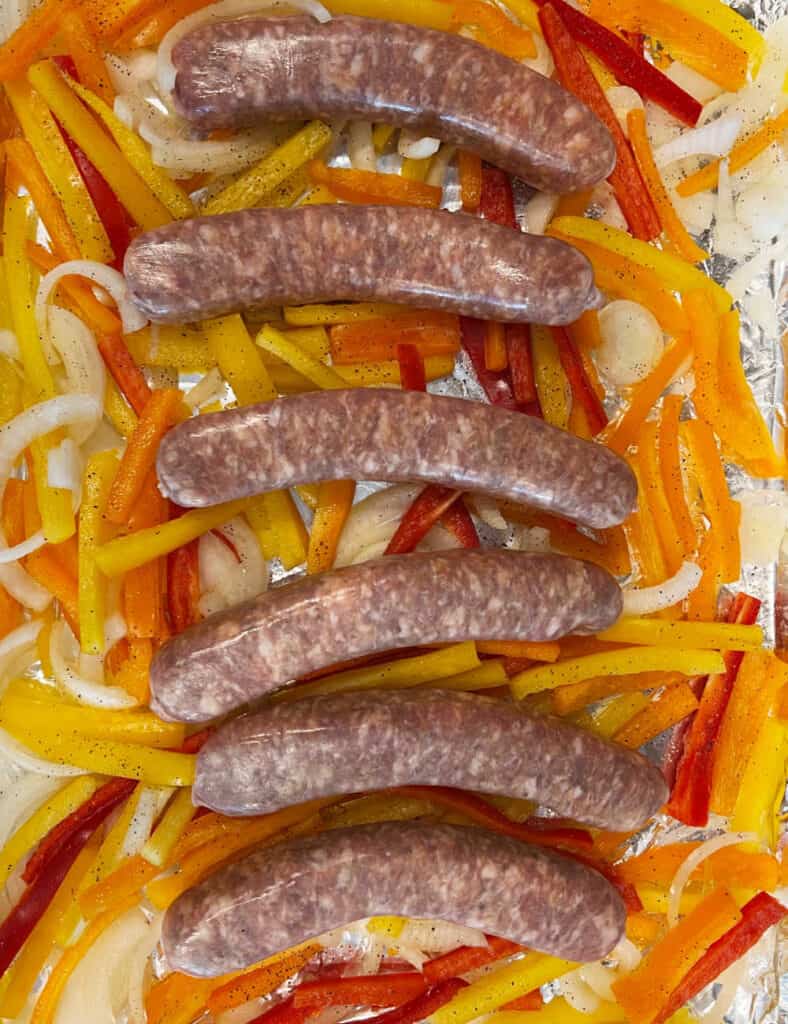 Italian sausages on top of sliced onions and peppers on baking sheet.