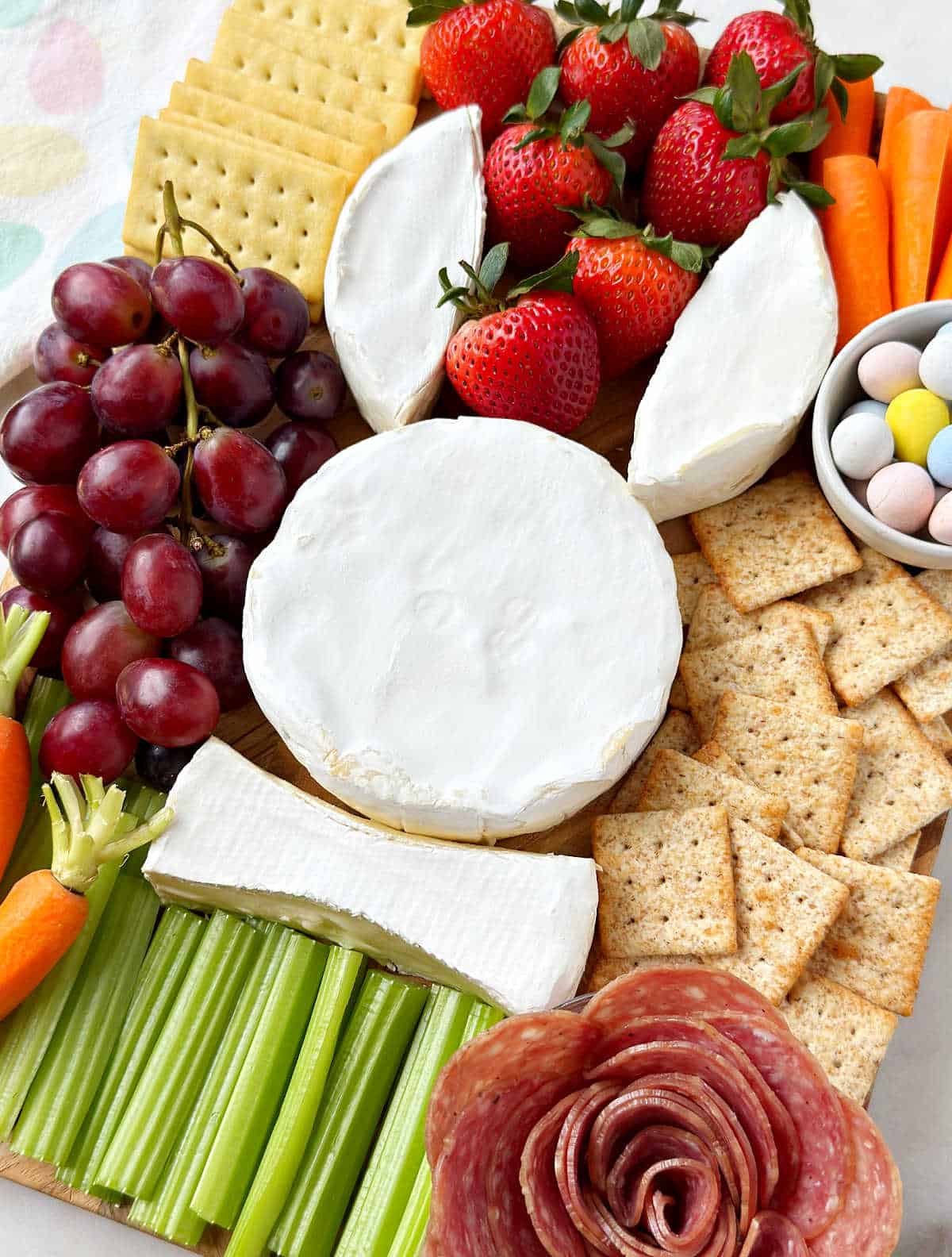 Easter charcuterie board with brie bunny, meat rose, veggies, fruit, candy eggs and crackers.