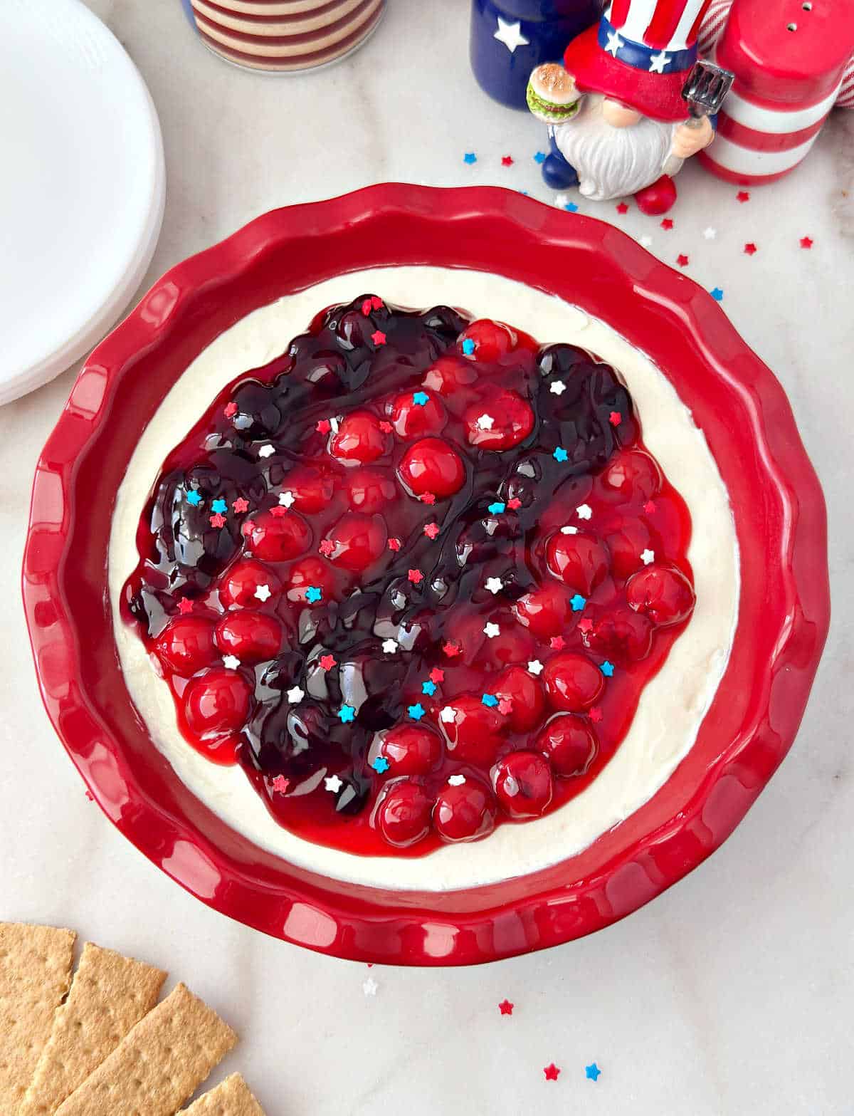 red white and blue cheesecake dip with cherries and blueberries in red pie dish.