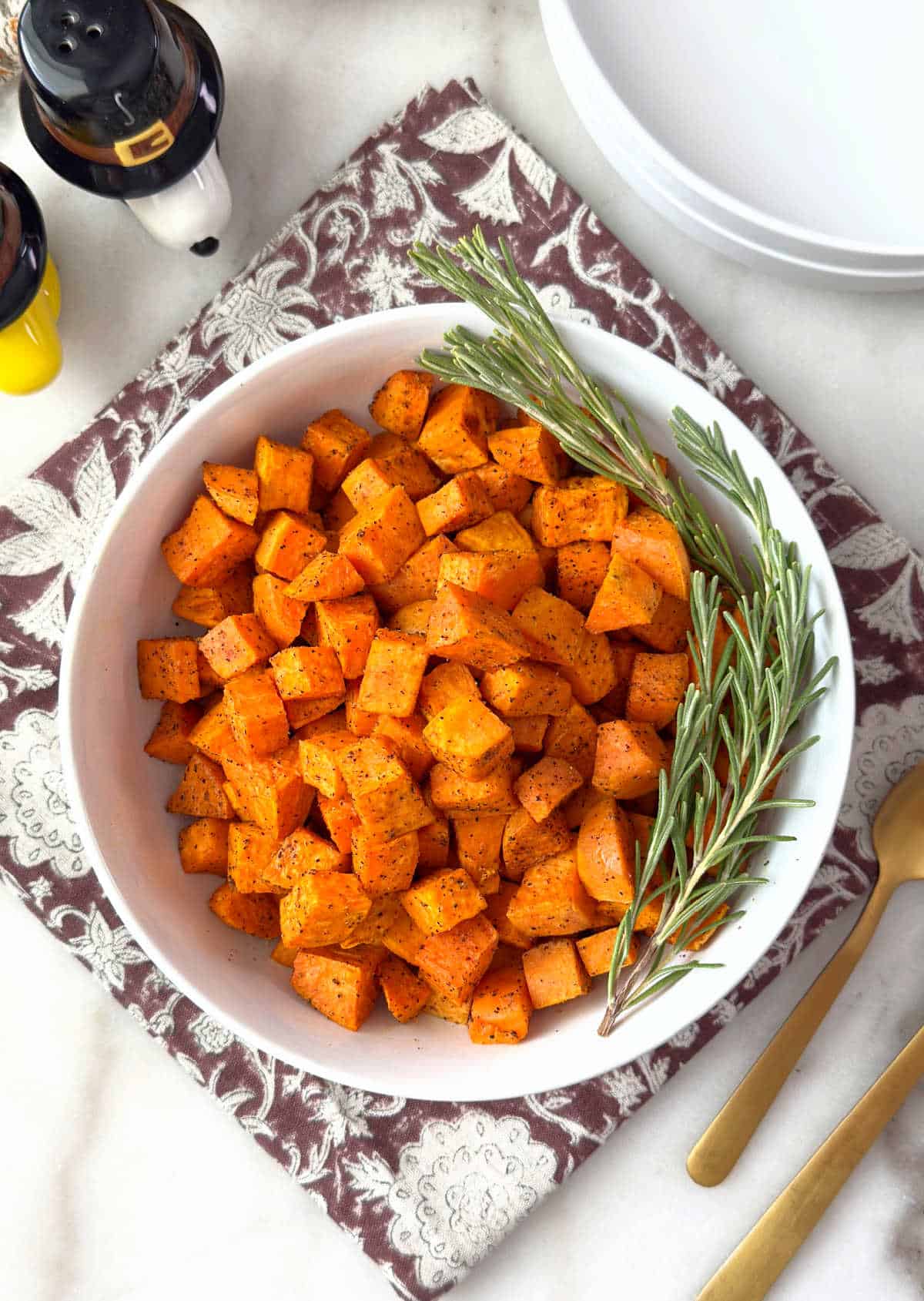 bowl of oven roasted sweet potatoes with rosemary sprigs on table.