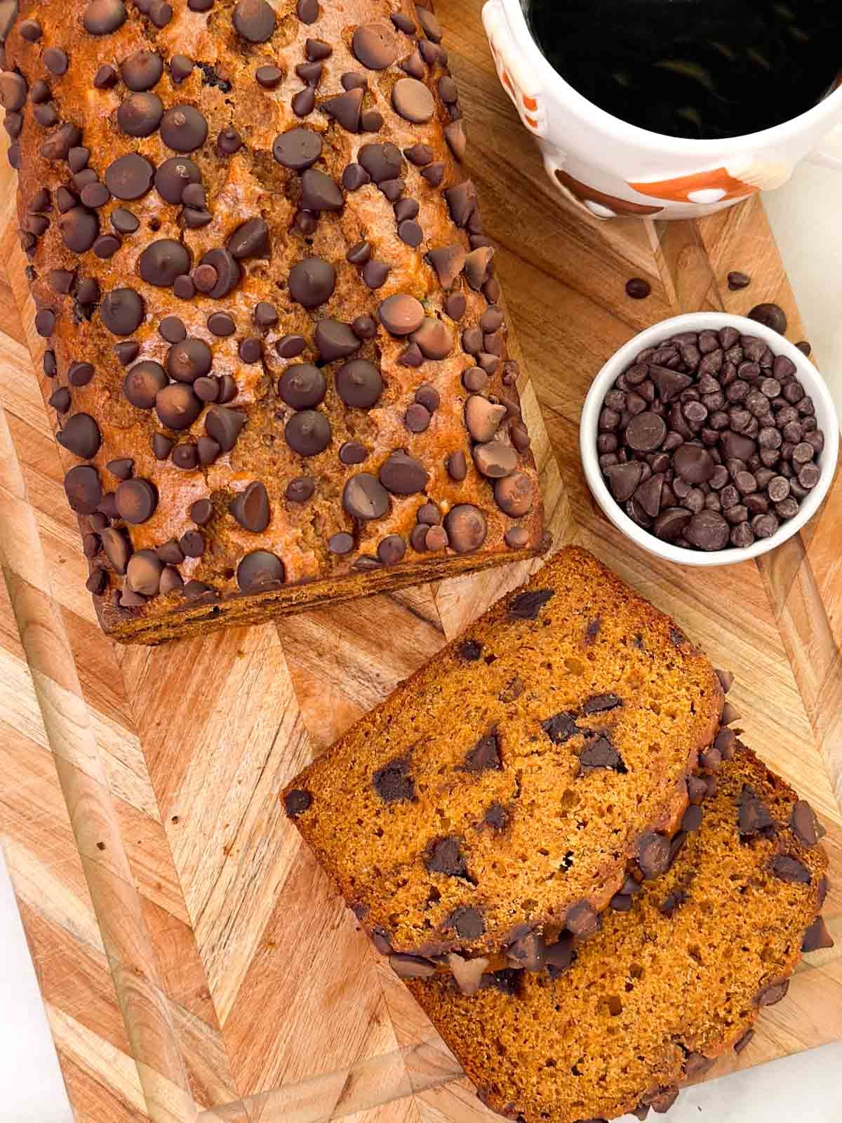 loaf of pumpkin banana bread with chocolate chips on cutting board with cup of coffee.